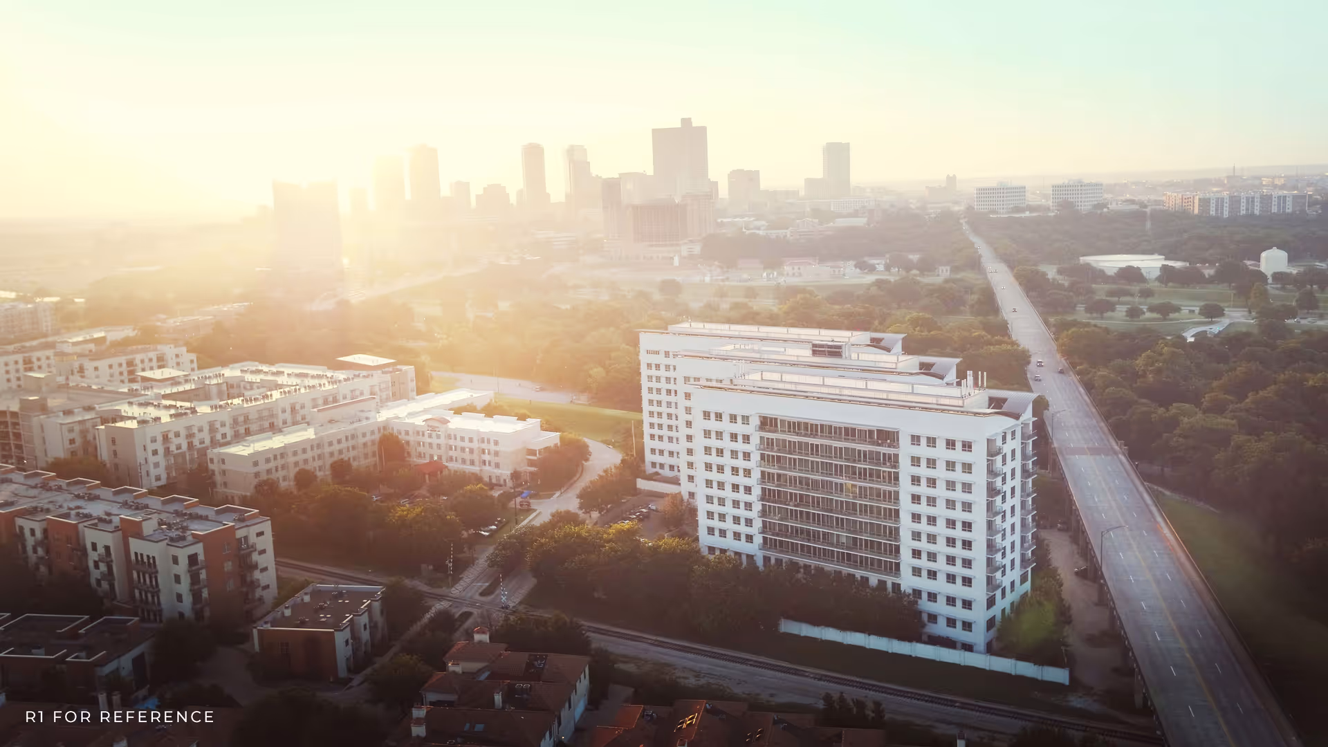 Aerial view of a large white multi-story residential building beside a long road with surrounding greenery and a hazy city skyline.