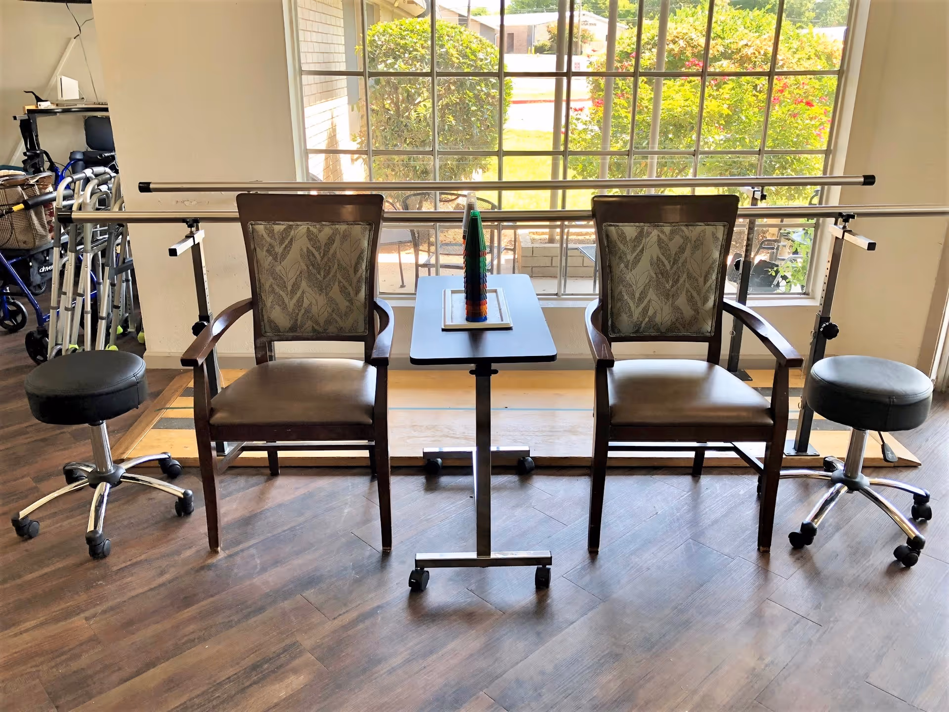 Two chairs and rolling stools beside parallel walking bars in a rehabilitation area by a large window.