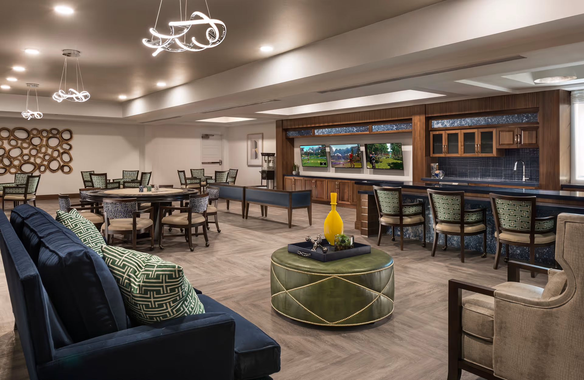 A spacious and modern common area with a mix of seating options including a dark blue sofa with green patterned pillows, round tables with chairs, a bar counter with high chairs, and a green ottoman with decorative items. The room features contemporary lighting fixtures, a wooden cabinet with three mounted flat-screen TVs, and a kitchenette area with a sink and glass-front cabinets.