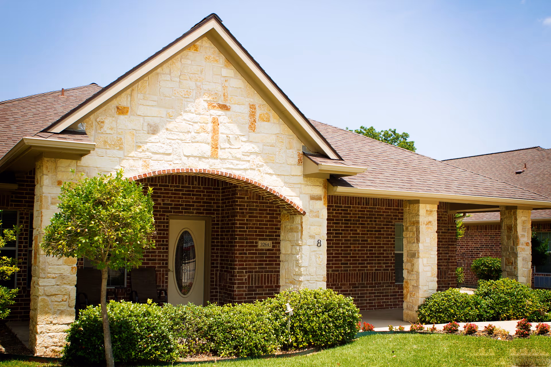 Exterior view of a single-story assisted living facility building with a brick and stone facade, a covered entrance with an arched stone detail, a door with an oval glass window, and well-maintained landscaping including bushes and a small tree under a clear blue sky.