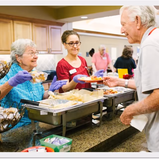 Two women wearing gloves serve food to an elderly man at a buffet-style counter in a kitchen or dining area. The women are smiling and handing a plate and a container of food to the man. Other people are visible in the background.