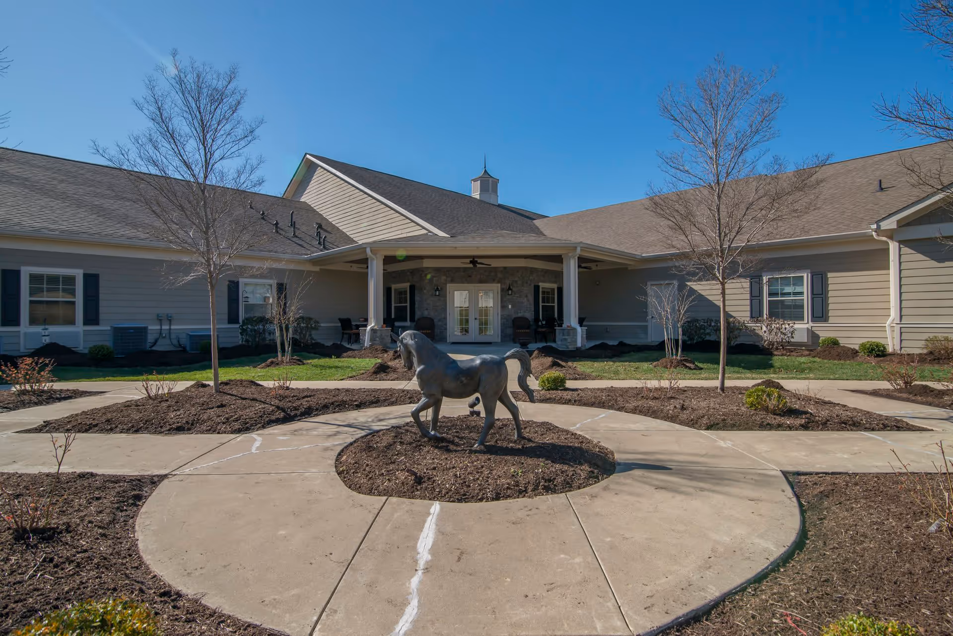 Exterior view of The Lantern at Morning Pointe Alzheimer's Center of Excellence in Frankfort showing a single-story building with a covered entrance, leafless trees, landscaped garden beds, and a bronze horse statue in the center of a circular concrete pathway.