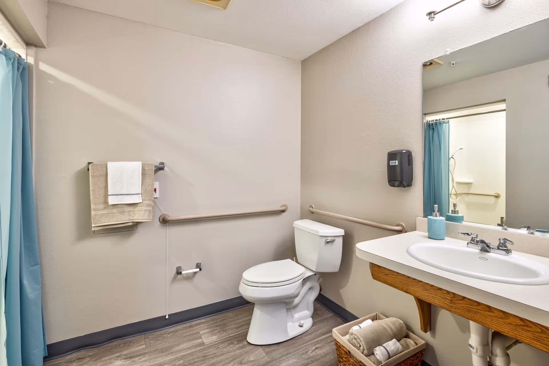 A clean and accessible bathroom with a white toilet, a sink with a large mirror above it, a soap dispenser mounted on the wall, and a basket with rolled towels underneath the sink. There are grab bars on the walls near the toilet and a towel rack with beige and white towels. A blue shower curtain is partially visible on the left side.