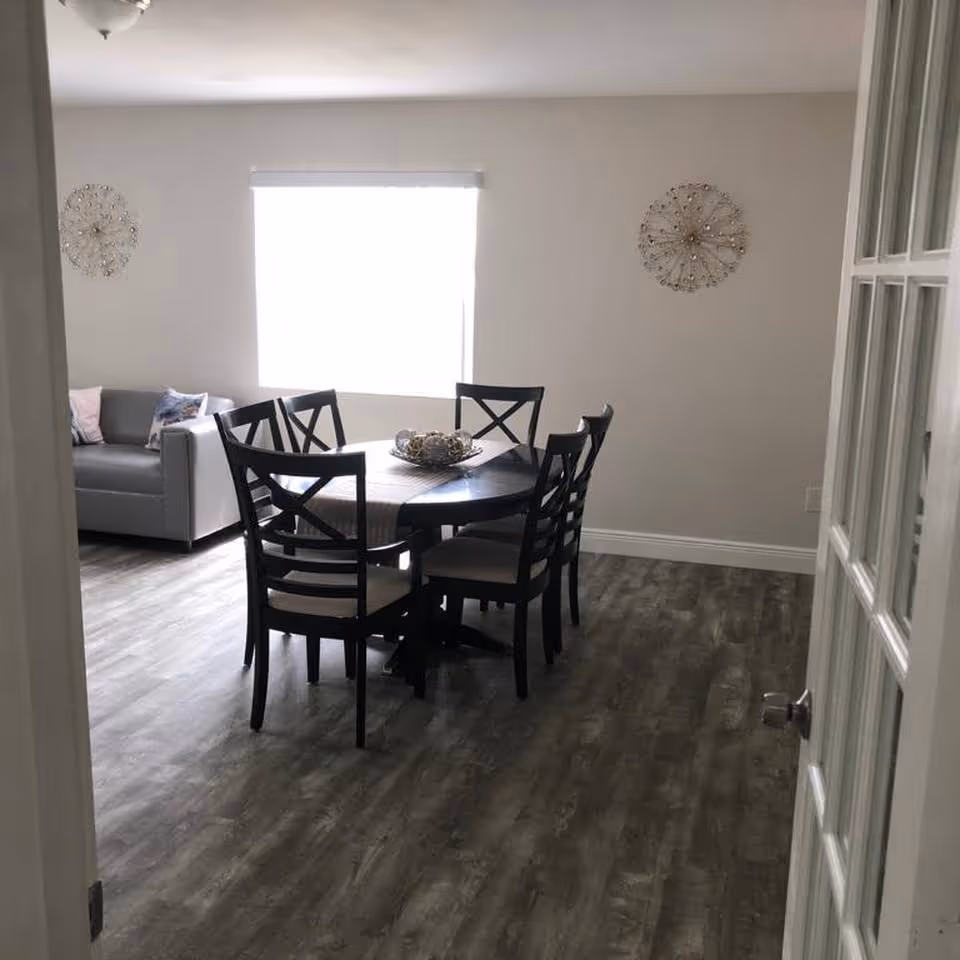 Interior view of a dining area with a round wooden table surrounded by six chairs with cushioned seats. A decorative centerpiece is placed on the table. In the background, there is a gray sofa with pillows against a wall with two decorative wall hangings. The floor has a wood-like finish and a window with a blind allows natural light into the room.