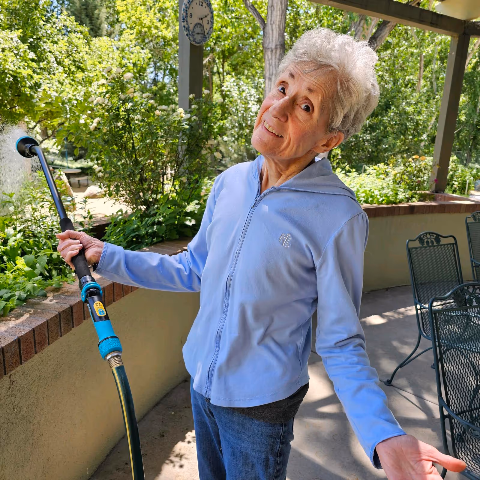 An elderly woman wearing a light blue zip-up jacket and jeans is standing outdoors on a patio, holding a garden hose with water spraying out. She is smiling and tilting her head slightly. The background shows green bushes, trees, a clock mounted on a post, and patio furniture.