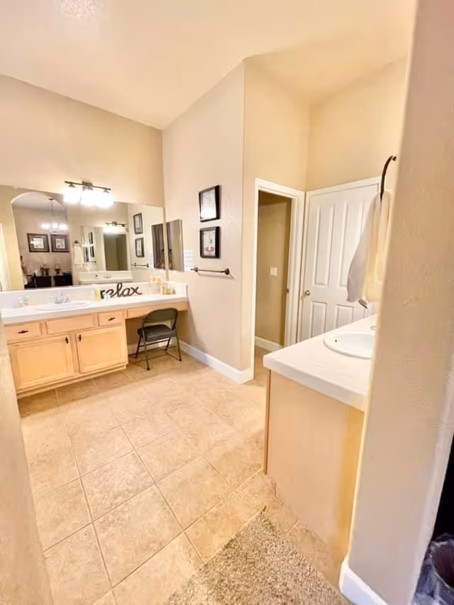 Interior view of a bathroom with beige tiled floor and light-colored walls. There is a large mirror above a wooden vanity with a sink and a small black folding chair underneath. On the vanity, there is a decorative sign that says 'relax'. To the right, there is another sink with a towel hanging on a ring. A door is visible in the background, and framed pictures hang on the wall.