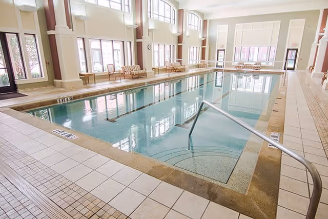 Indoor swimming pool with clear water surrounded by tiled flooring and large windows letting in natural light. Several chairs and tables are placed along the far side of the pool area.