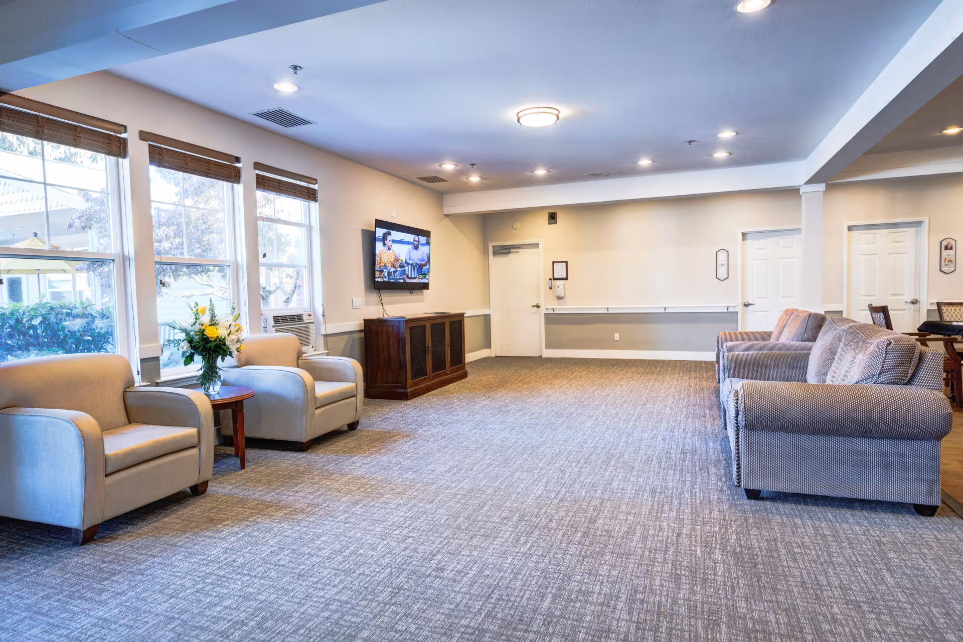 A spacious and well-lit living room area with large windows on the left side, two beige armchairs with a small round wooden table holding a vase of flowers between them, and three gray upholstered sofas on the right side. A flat-screen TV is mounted on the wall above a wooden cabinet. The room has a carpeted floor and recessed ceiling lights.