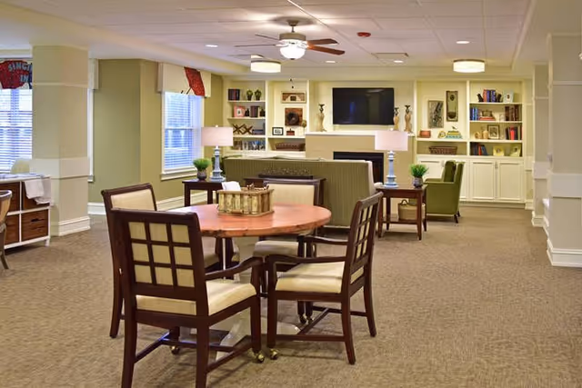 A furnished senior living common room with a round table and chairs in the foreground and sofas, armchairs, a wall-mounted TV and built-in bookshelves in the background.