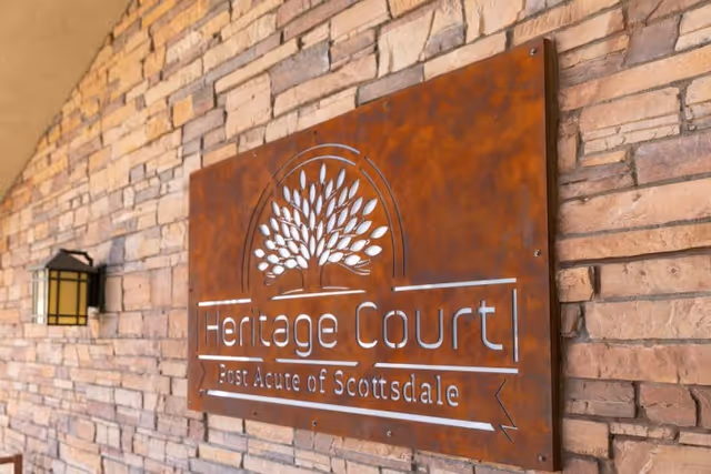 A rust-colored metal sign mounted on a stone wall that reads 'Heritage Court Post Acute of Scottsdale' with a tree logo above the text. A black and yellow outdoor wall lantern is visible to the left of the sign.