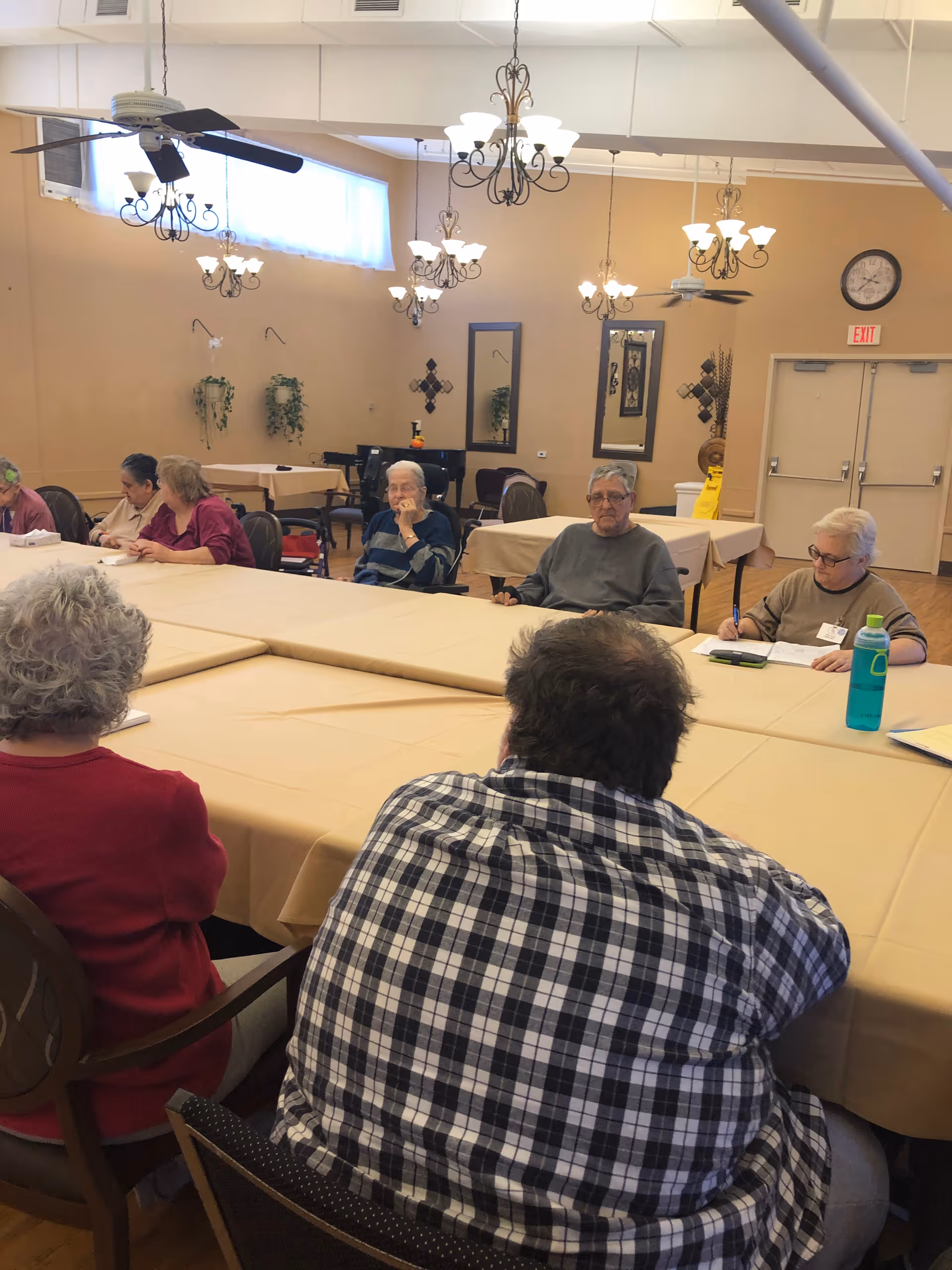 A group of elderly people sitting around tables covered with beige tablecloths in a well-lit room with chandeliers and ceiling fans. The room has beige walls with decorative mirrors and plants, and a clock and exit sign are visible above double doors in the background.