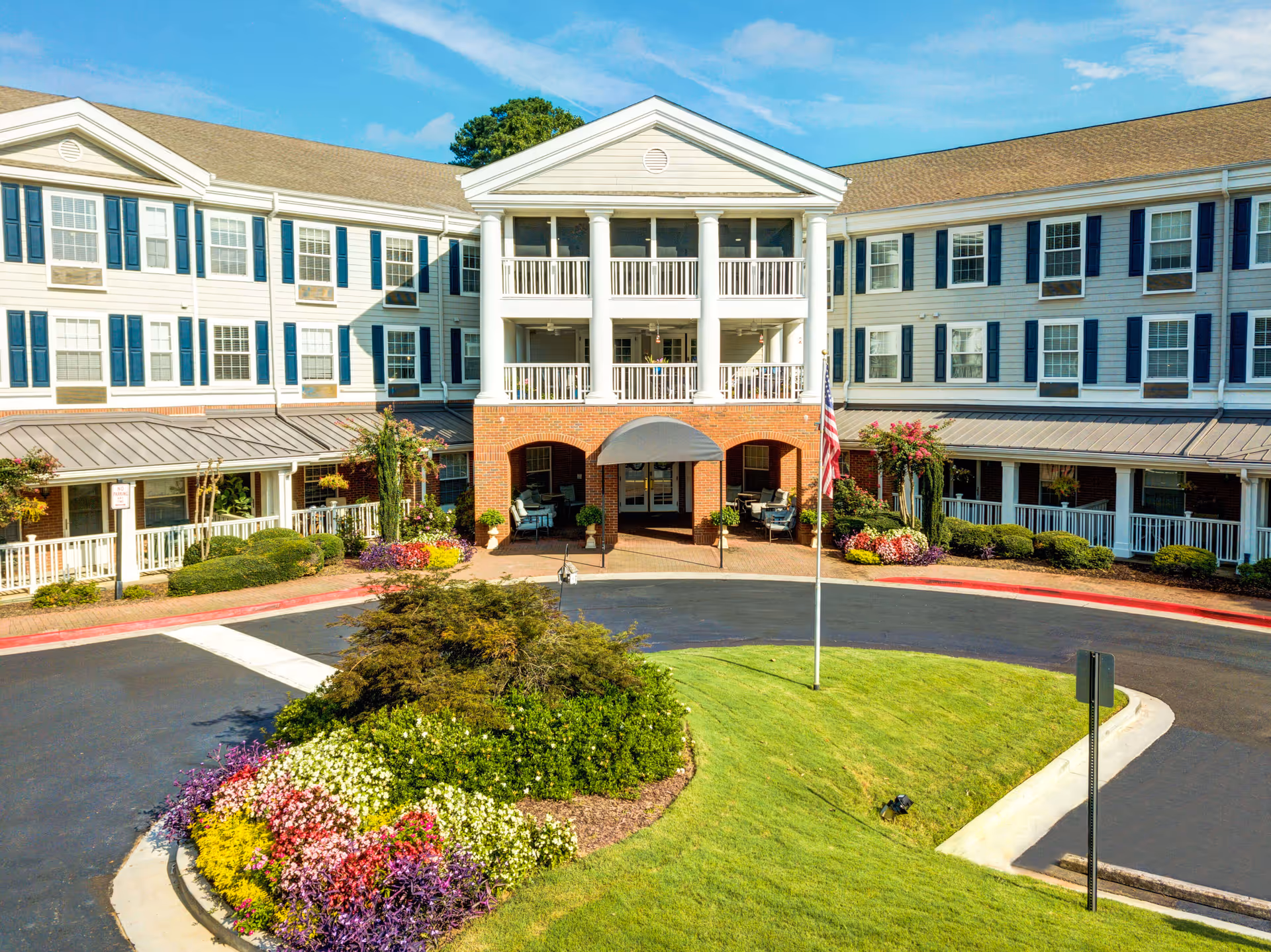 Front exterior view of a large senior living facility named Historic Roswell Place, featuring a three-story building with white siding, blue shutters, and a brick entrance with white columns. The driveway circles a landscaped area with colorful flowers and greenery, and an American flag is displayed on a flagpole in front.