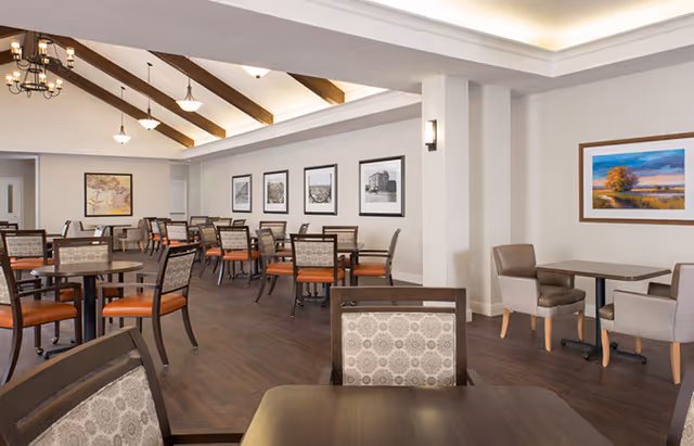 A spacious dining room with multiple tables and chairs arranged neatly. The room features wooden floors, white walls adorned with framed artwork, and a ceiling with exposed wooden beams and hanging light fixtures. There are cushioned chairs with patterned upholstery and some with solid orange seats.