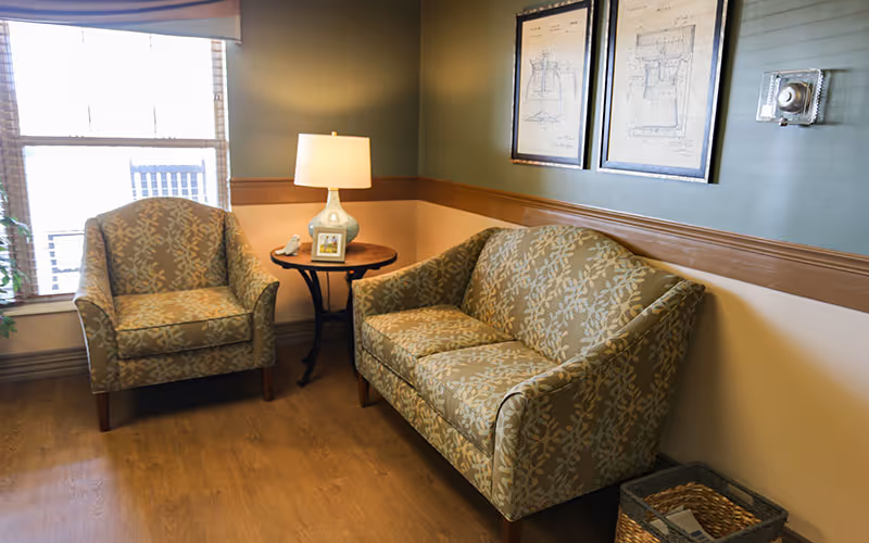 Small lounge with two patterned armchairs and a loveseat, a round side table with a lamp, framed prints on the wall, and a window.