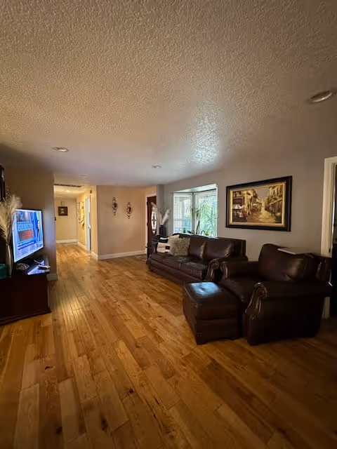 Interior view of a living room in Sunnyside Adult Care Home featuring wooden flooring, a brown leather sofa with cushions, a matching leather armchair with an ottoman, a framed painting on the wall, a window with blinds, and a television on a stand to the left. The room extends into a hallway with light-colored walls and ceiling lights.
