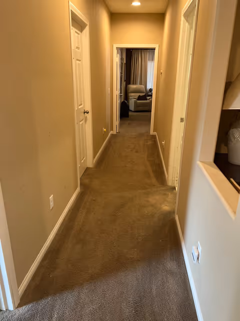 Carpeted hallway with beige walls and doors leading to a living area with an armchair at the end.