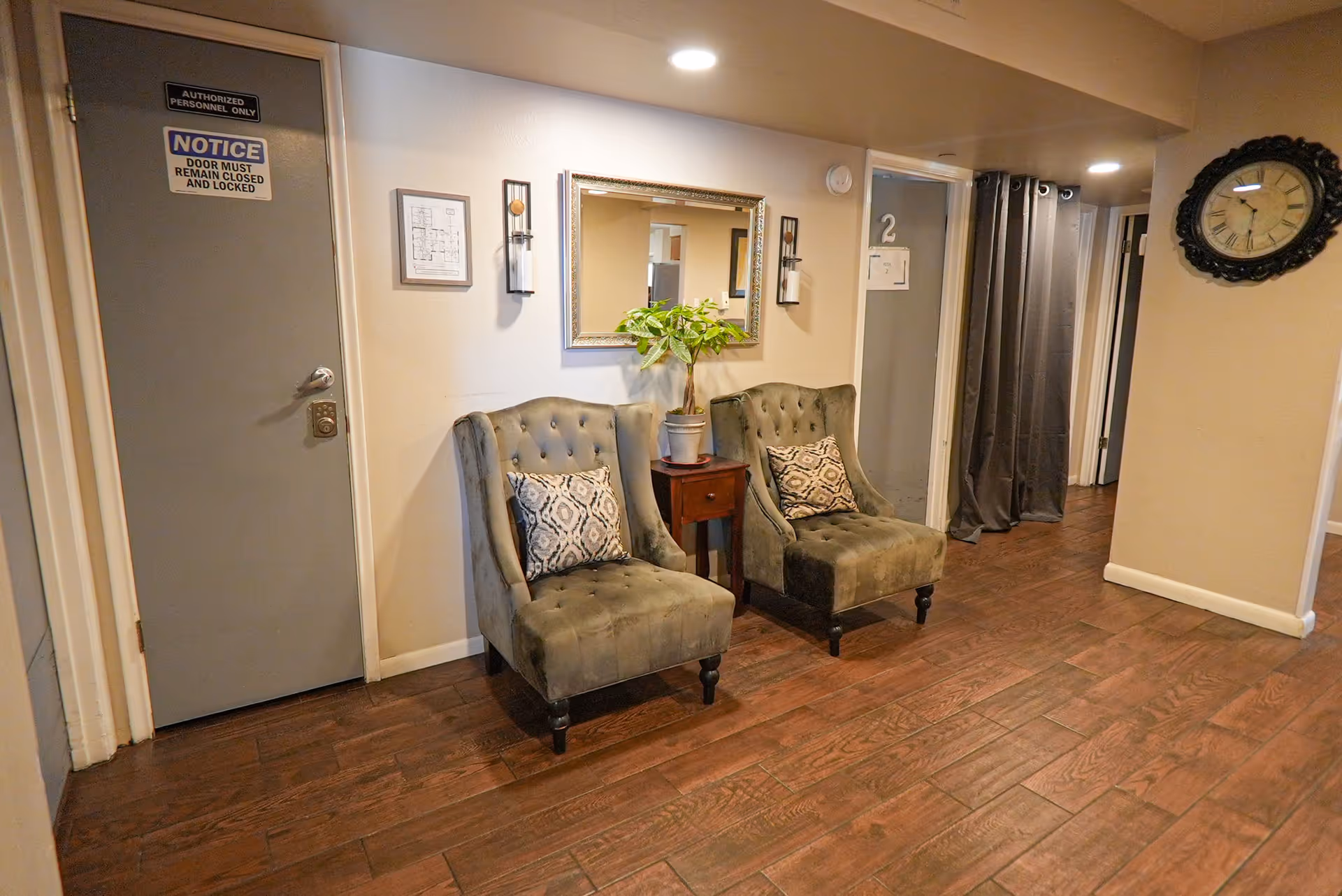 A hallway in an assisted living facility with two gray upholstered chairs with patterned cushions, a small wooden side table with a potted plant, a large wall mirror, and a clock on the wall. The floor is wooden, and there are doors and a curtain visible in the background.