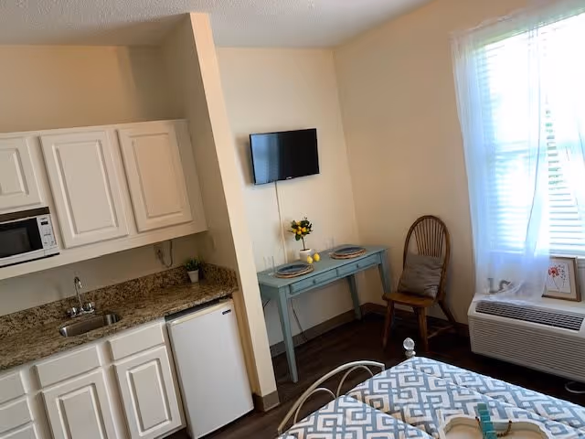 A small senior living studio room featuring a kitchenette with white cabinets, a microwave, a sink, and a mini refrigerator. Adjacent to the kitchenette is a small blue table set with two plates and a vase with yellow flowers, accompanied by a wooden chair with a cushion. A flat-screen TV is mounted on the wall above the table. The room has a window with sheer white curtains and a heating/cooling unit below it. Part of a bed with a patterned blue and white bedspread is visible in the foreground.