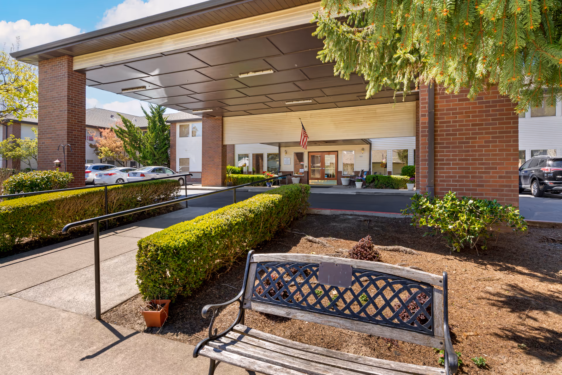 Entrance of Brookdale McMinnville City Center showing a covered drop-off area with brick pillars, a ramp with handrails, trimmed hedges, a wooden bench, and an American flag hanging near the glass doors of the building.