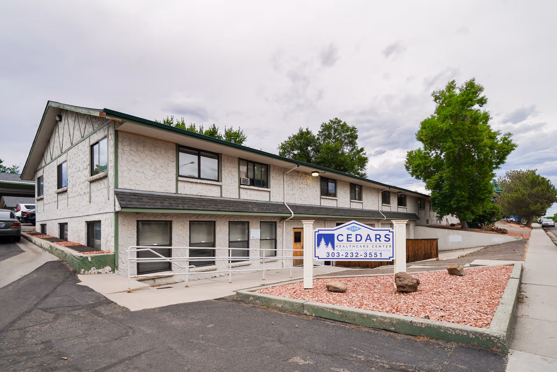 Exterior view of Cedars Health Care Center, a two-story building with textured white walls and green trim. There is a sign in front displaying the facility's name and phone number. The area around the building includes a paved driveway, a sidewalk, some landscaping with rocks, and several trees under a cloudy sky.