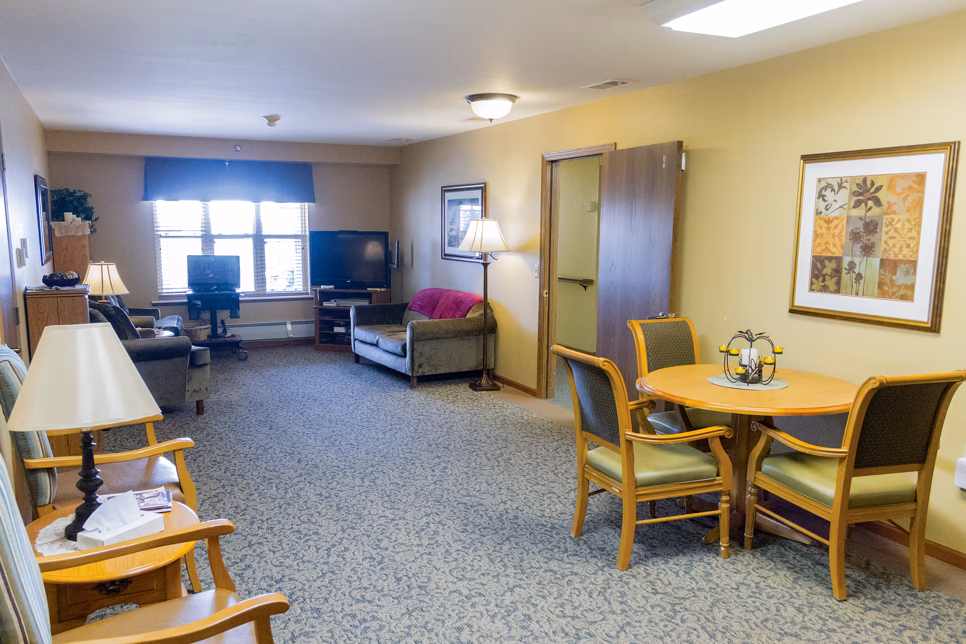 A senior living facility common area with beige walls and carpeted floor. The room features a round wooden table with four chairs on the right side, a framed floral artwork on the wall above the table, and a floor lamp next to a gray loveseat with a red throw blanket. At the far end, there is a window with blinds and two televisions on stands. On the left side, there are two wooden armchairs with cushions and a small table with a lamp and tissue box.