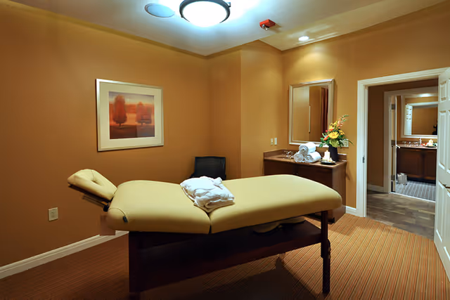 A warmly lit room with a massage table covered in a beige sheet and a folded white towel on top. The room has brown walls, a framed picture of trees, a black chair, a wooden cabinet with a mirror above it, and a vase with flowers. An open door reveals a glimpse of an adjoining room with a sink and mirror.