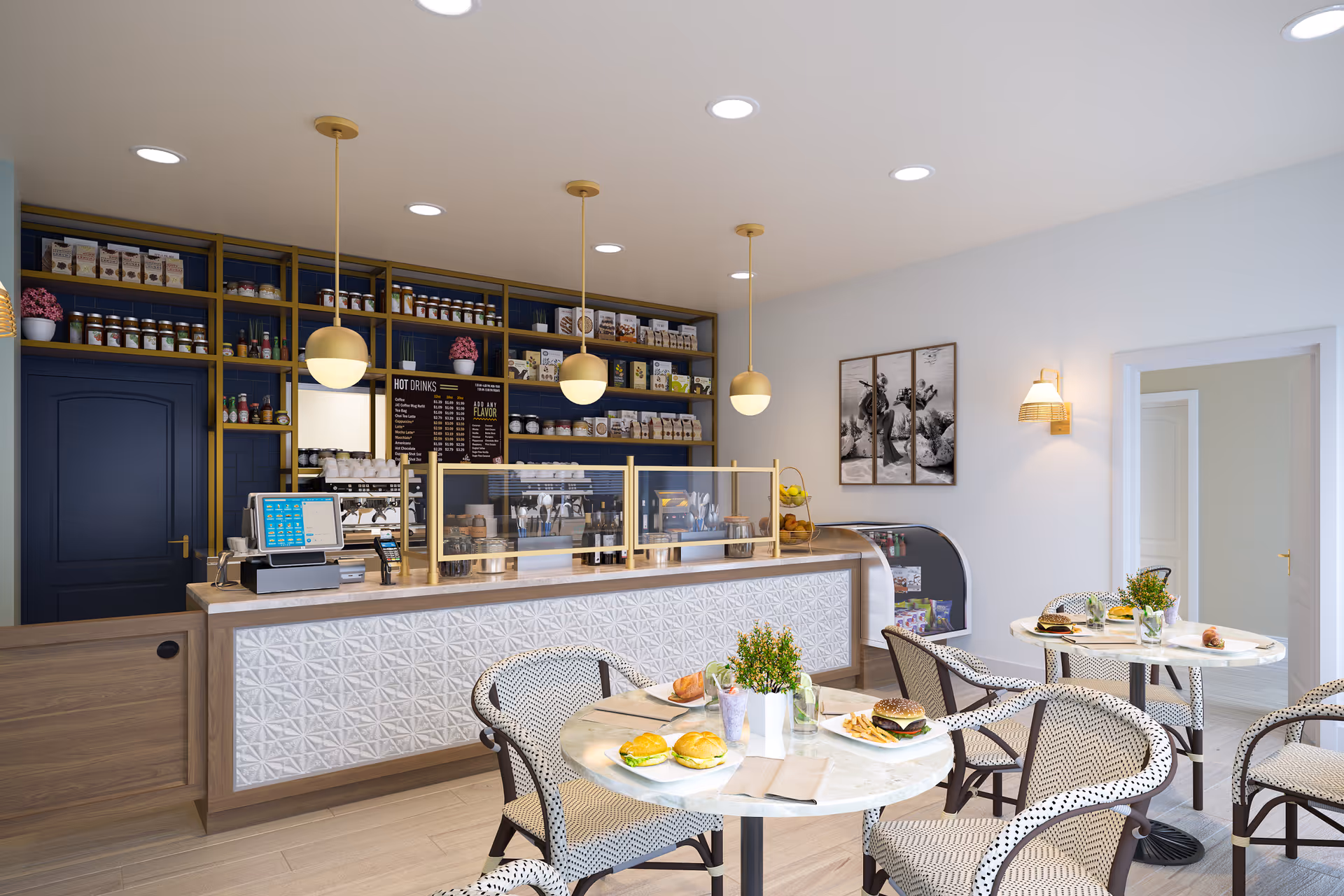 Interior view of a modern cafe area with a counter displaying various jars and packaged goods on shelves behind it. There are three round tables with patterned chairs, each table set with plates of food including sandwiches, a burger with fries, and drinks. The space is well-lit with ceiling lights and hanging pendant lamps, and there is a small refrigerated display case near the wall with framed black and white artwork above it.