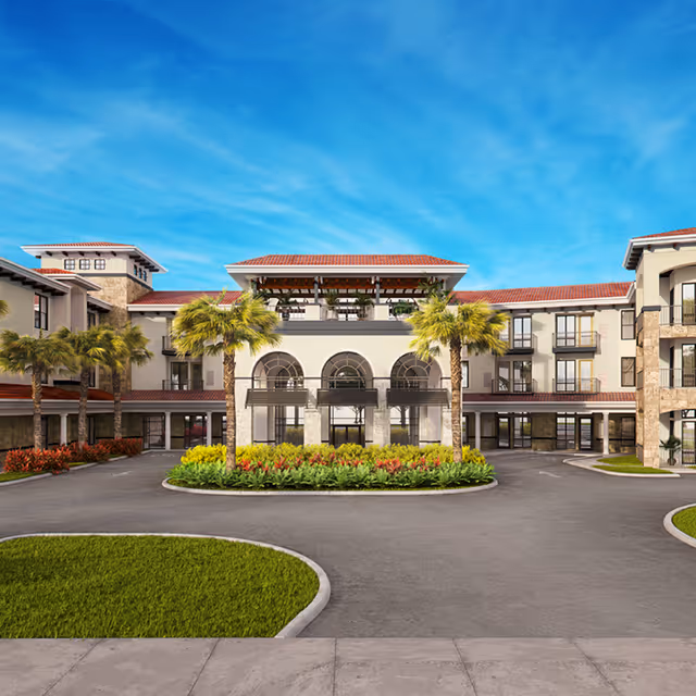 Front exterior view of Heartis Venice Assisted Living facility featuring a large, multi-story building with arched windows, red-tiled roofs, palm trees, and landscaped greenery under a bright blue sky.