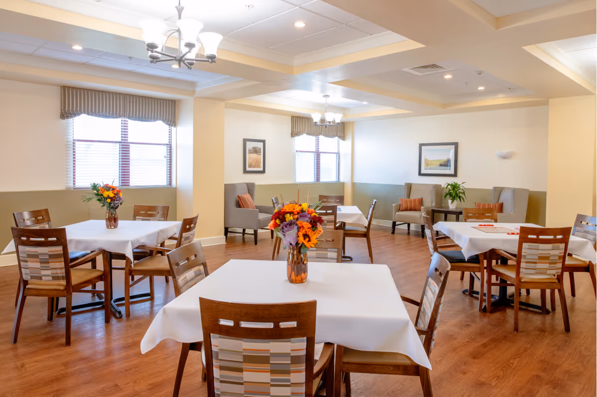 A bright and clean dining room with several wooden tables covered with white tablecloths. Each table has a vase with colorful flowers. The room has wooden flooring, beige walls with a green lower half, and large windows with striped valances. There are comfortable armchairs with orange cushions along the walls and framed pictures hanging above them. Ceiling lights provide additional illumination.