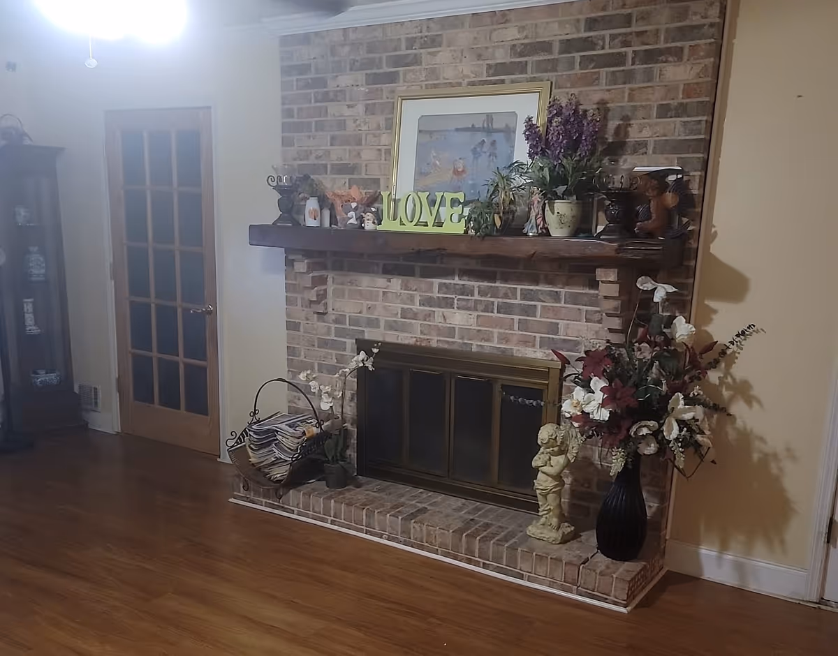A cozy living room area featuring a brick fireplace with a wooden mantel decorated with plants, a framed picture, and a decorative 'LOVE' sign. To the right of the fireplace is a large vase with artificial flowers and a small statue. To the left is a basket with magazines and a small potted plant. The room has wooden flooring and a glass-paneled wooden door on the left side.