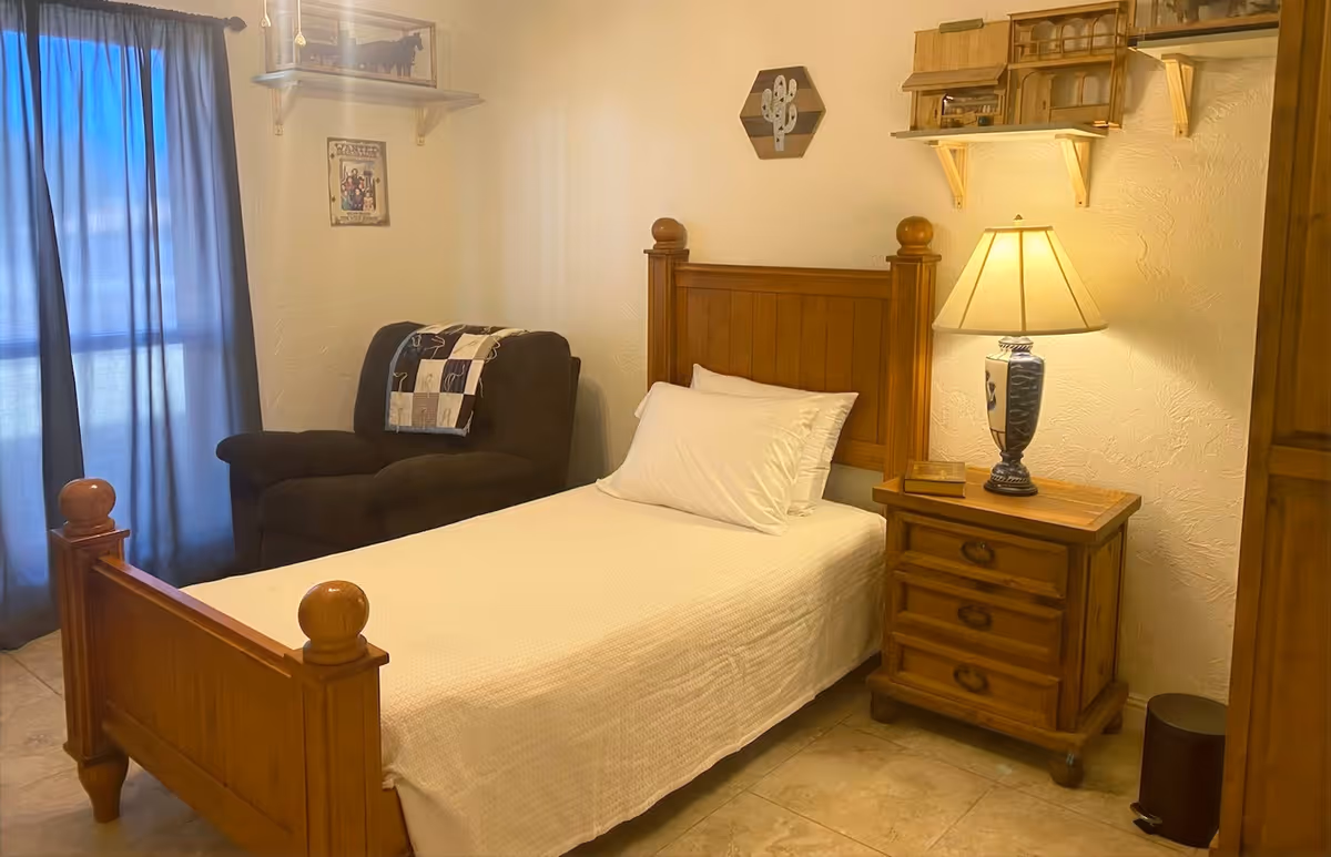 A cozy single bedroom with a wooden bed, nightstand and lamp, an armchair by blue curtains, and wall shelves.