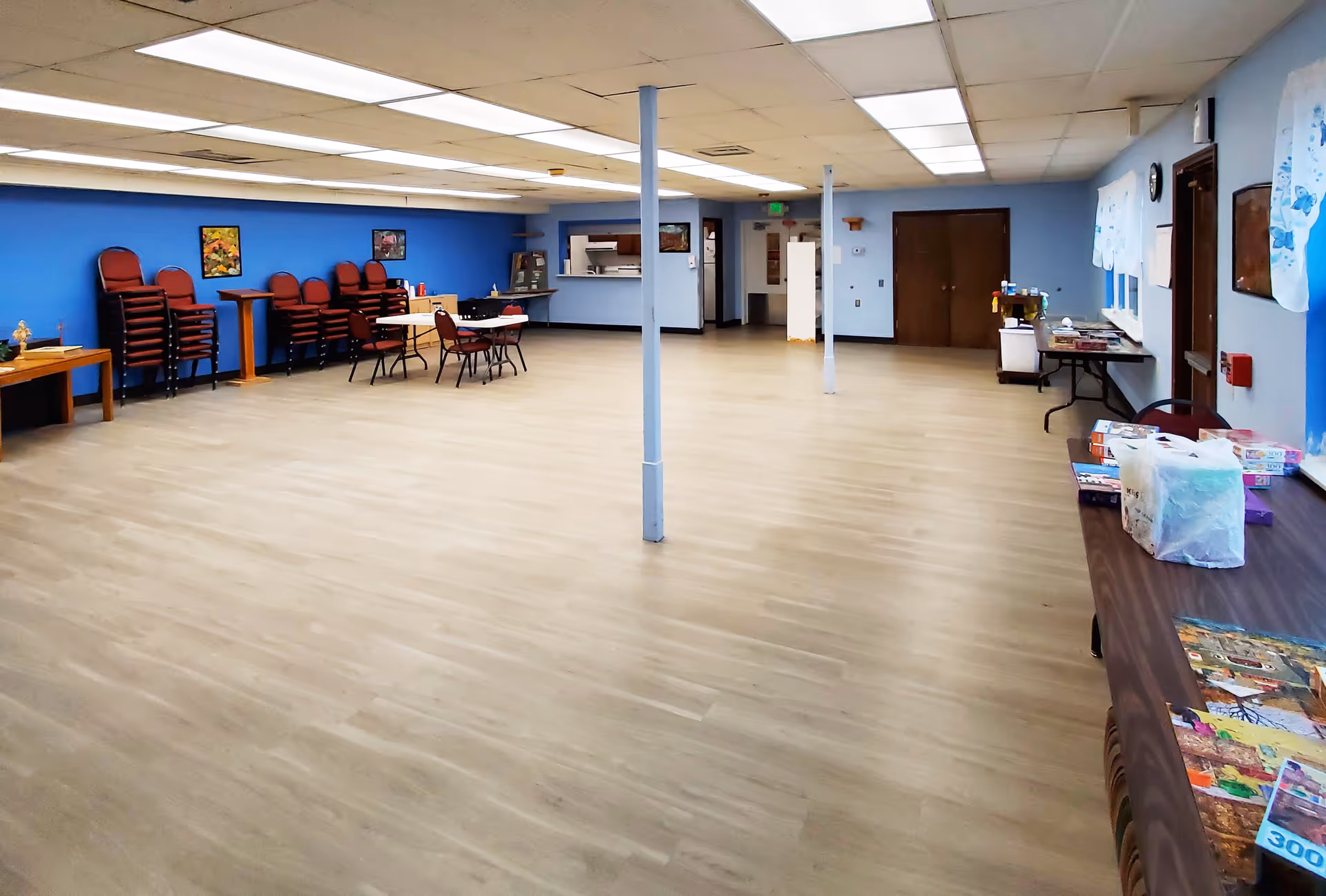 A large multipurpose room with light wood flooring and blue walls. The room contains stacked chairs, a few tables with chairs, and various items on tables along the right wall. There are two support columns in the middle of the room, fluorescent ceiling lights, and a small kitchen or serving window at the back.