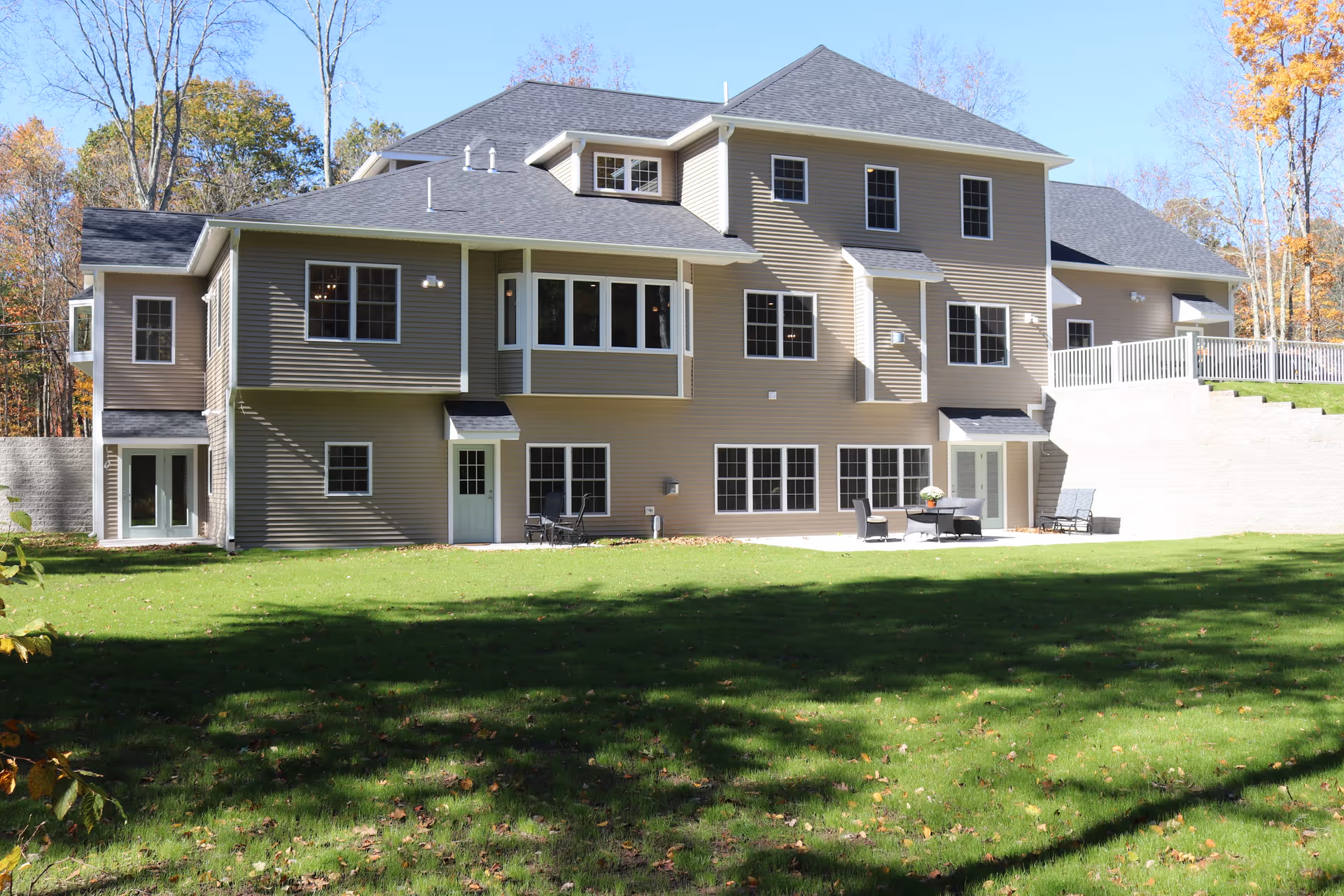 Rear exterior of a large two-story beige residential care home with many windows, a patio with chairs, and a grassy lawn backed by trees.