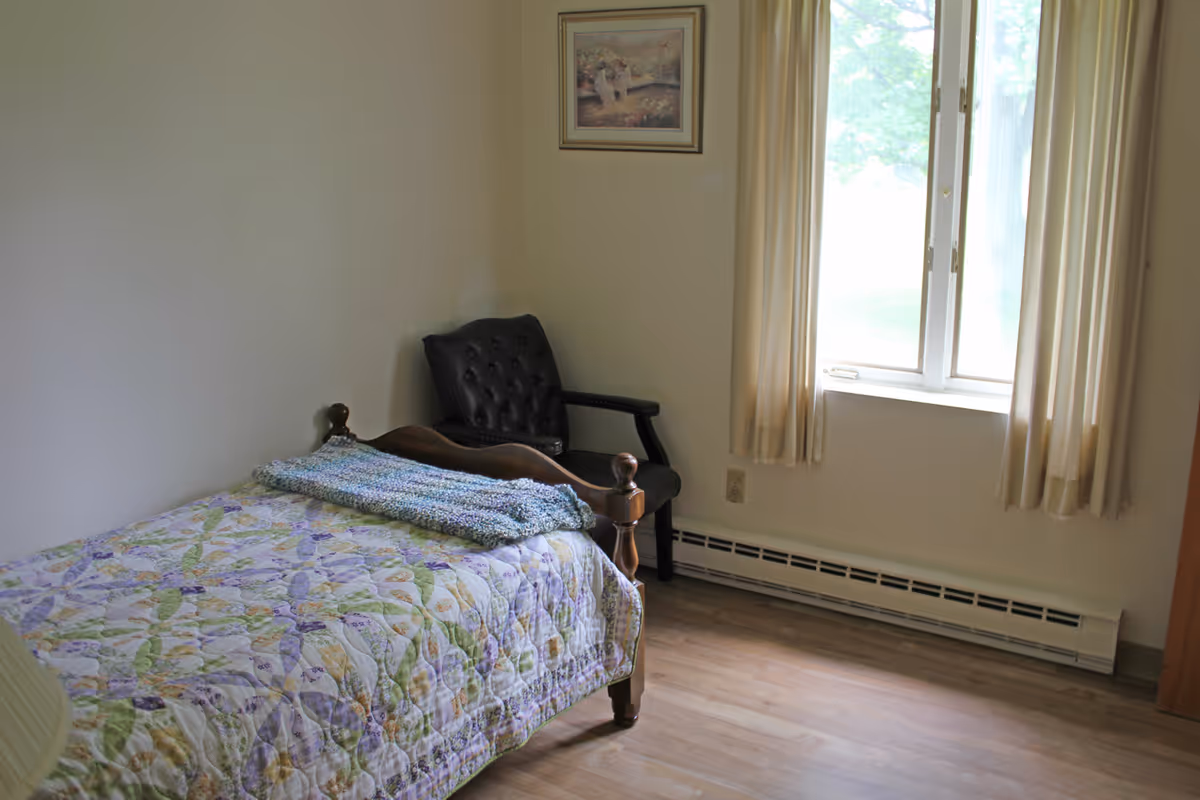 A simple bedroom with a single wooden bed covered with a floral quilt and a folded knitted blanket on top. Next to the bed is a black cushioned chair. A window with beige curtains allows natural light to brighten the room. A framed picture hangs on the wall above the chair.