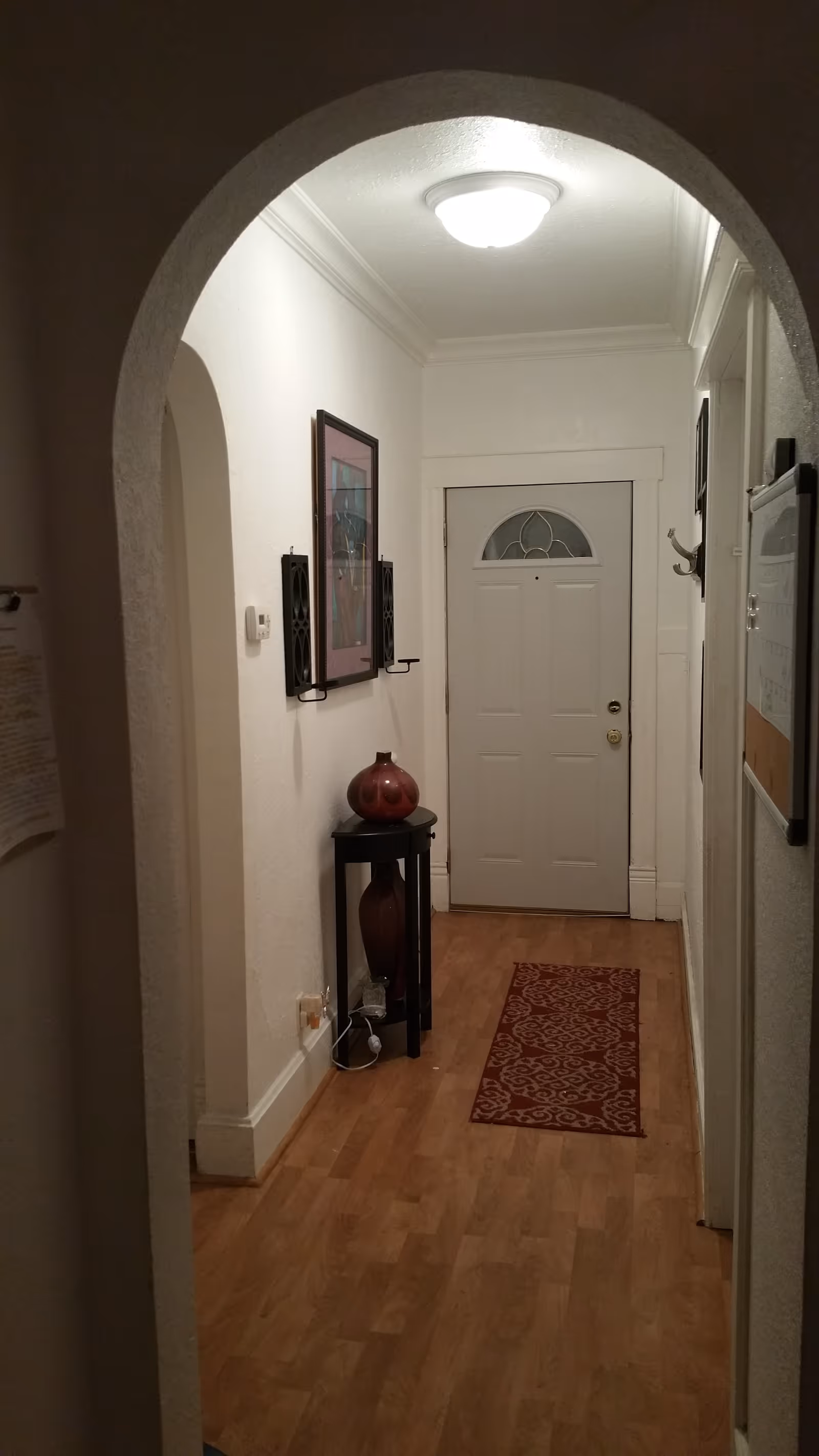 View through an arched doorway into a hallway with a closed white front door at the end. The hallway has light wood flooring and a red patterned runner rug. On the left wall, there is a framed picture and two black wall-mounted candle holders. Below them is a small black table with two decorative vases. The right wall has a coat hook and a bulletin board. A ceiling light fixture illuminates the space.
