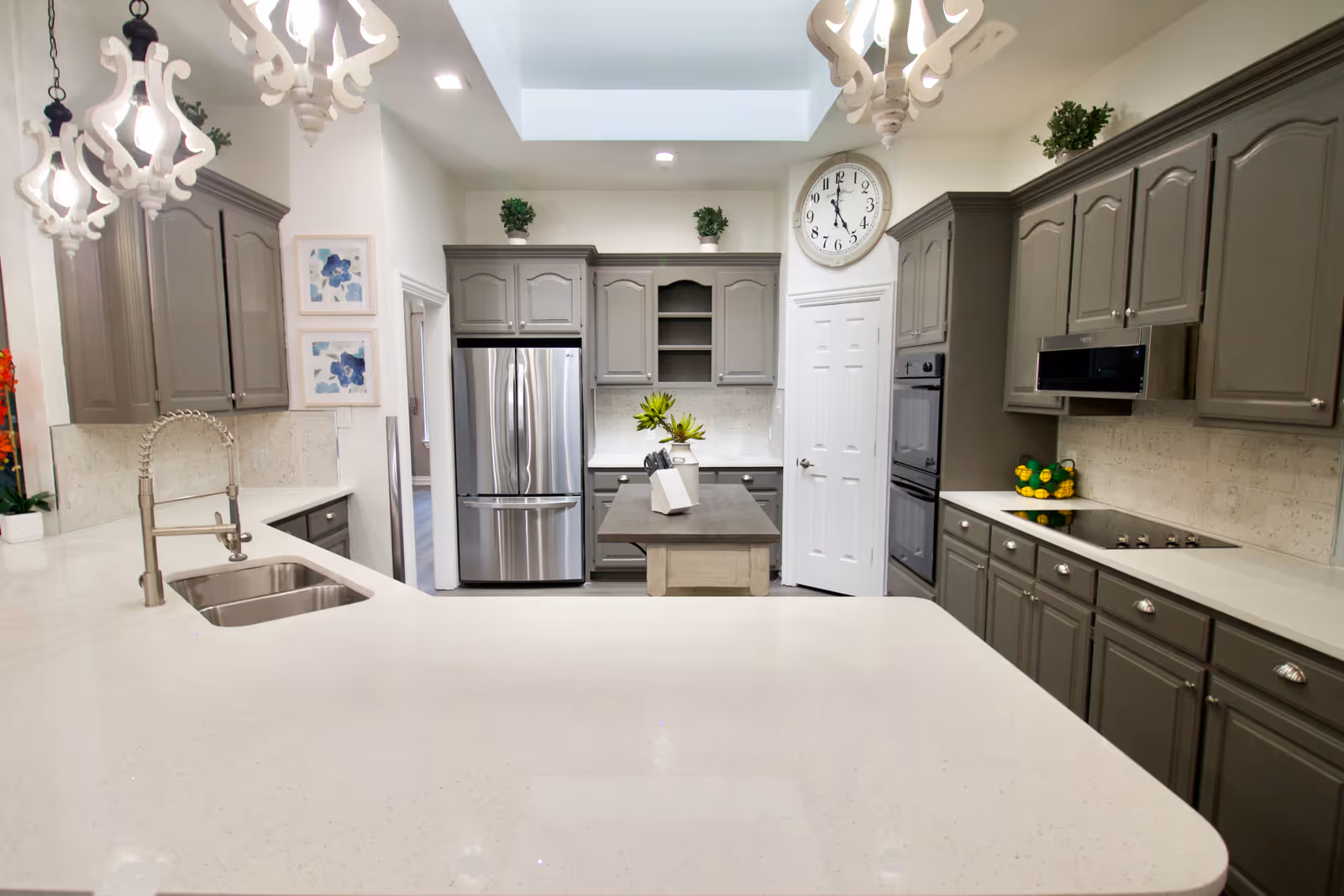 A modern kitchen with gray cabinets, a large white countertop island with a built-in sink and faucet, stainless steel refrigerator, double oven, and stovetop. The kitchen is well-lit with decorative hanging lights, a large wall clock, and small potted plants on top of the cabinets. There are also two framed floral artworks on the wall.