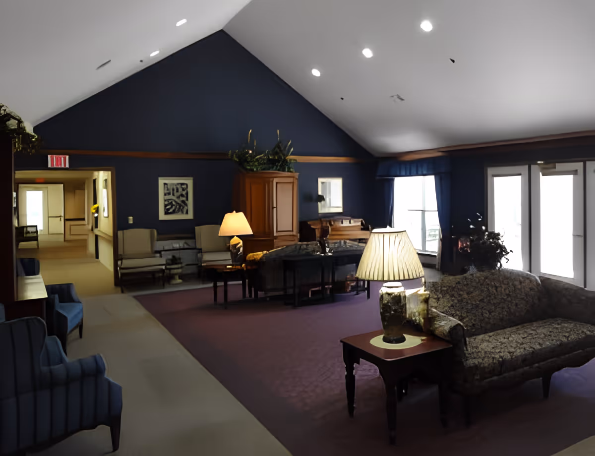 Interior view of a senior living facility lounge area with a high vaulted ceiling, blue walls, and carpeted floor. The room is furnished with various chairs, sofas, side tables with lamps, a wooden cabinet, and a piano. Large windows and glass doors allow natural light to enter the space.