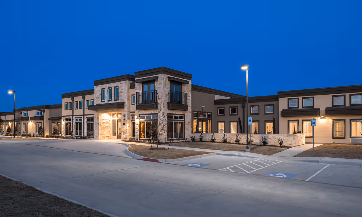 Exterior view of The Heritage at Twin Creeks facility at dusk, showing a modern building with stone and stucco facade, large windows, balconies, and illuminated entrance. The parking area in front includes handicapped parking spaces and street lamps.