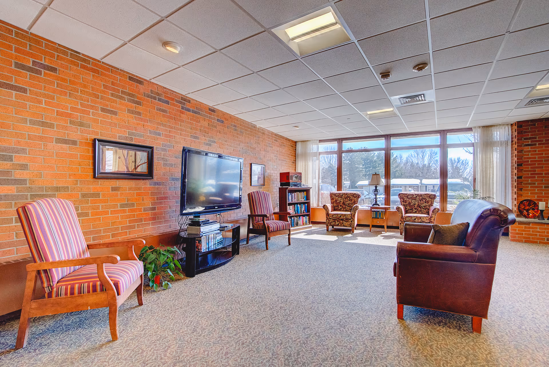 A bright and spacious living room area with a brick wall, a large flat-screen TV mounted on a stand, several upholstered chairs including two striped armchairs and two floral armchairs, a leather sofa, a small bookshelf, and large windows letting in natural light.