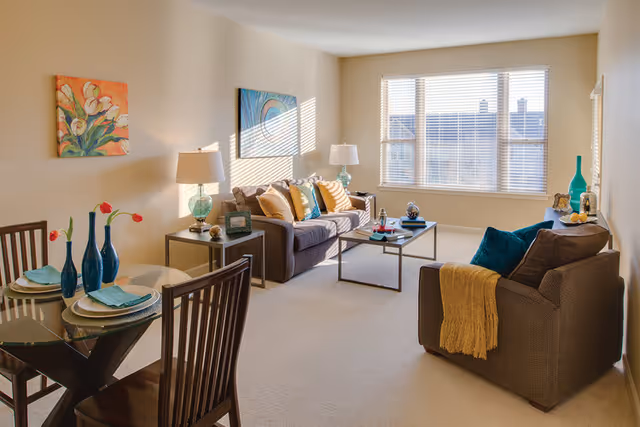 Bright furnished living room with a sofa, armchair, coffee table, dining table, and a large sunlit window.