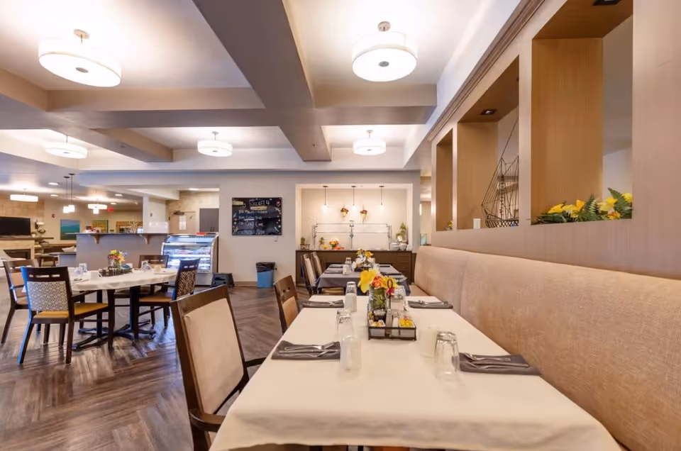 A bright and spacious dining room in a senior living facility with tables set for meals. The tables have white tablecloths, napkins, glasses, and small flower arrangements. The room features modern ceiling lights, wooden flooring, and a beige cushioned bench along one wall with decorative shelves above it.