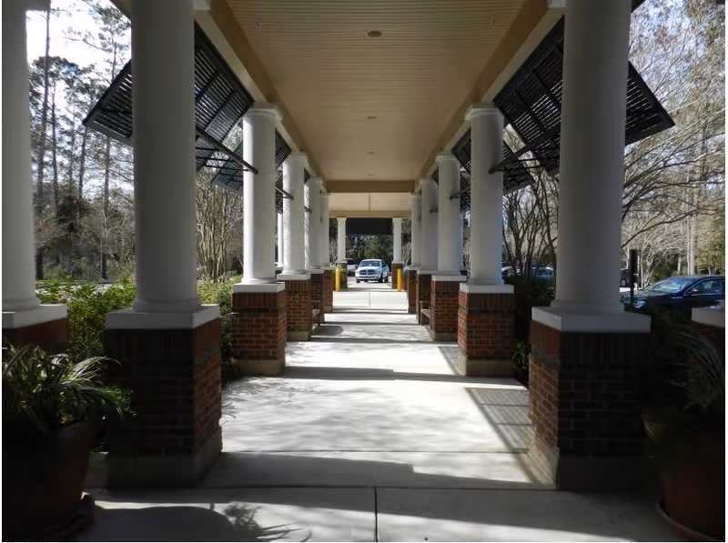 Covered walkway with white columns and brick bases leading to a parking area with cars visible in the background, surrounded by trees and greenery.