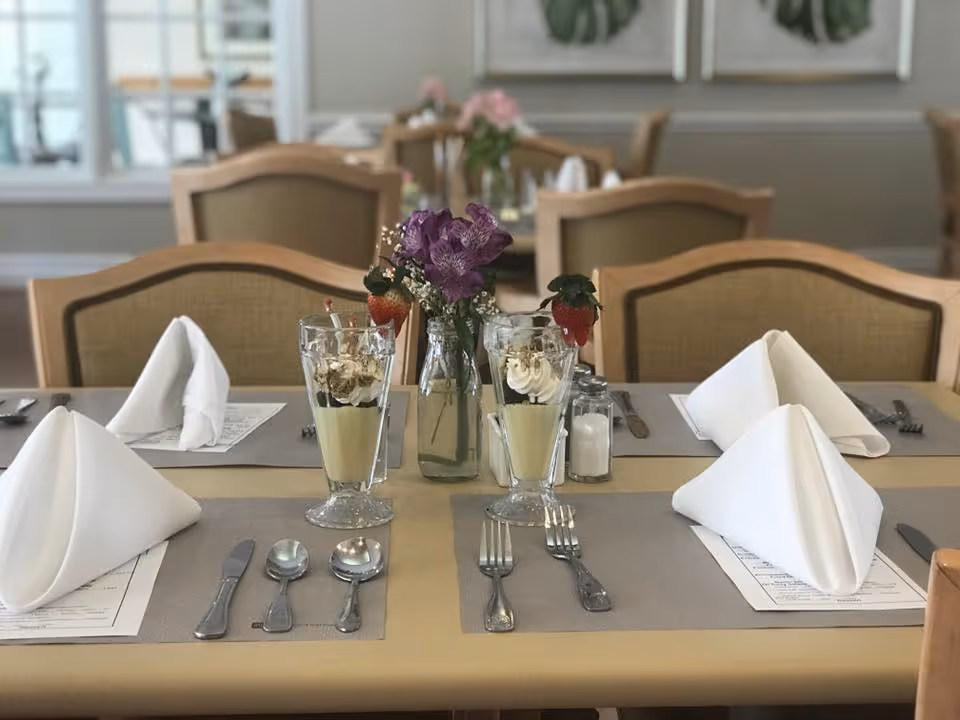 A dining table set with folded napkins, utensils, dessert parfaits with strawberries, and a small vase of flowers in a dining room.