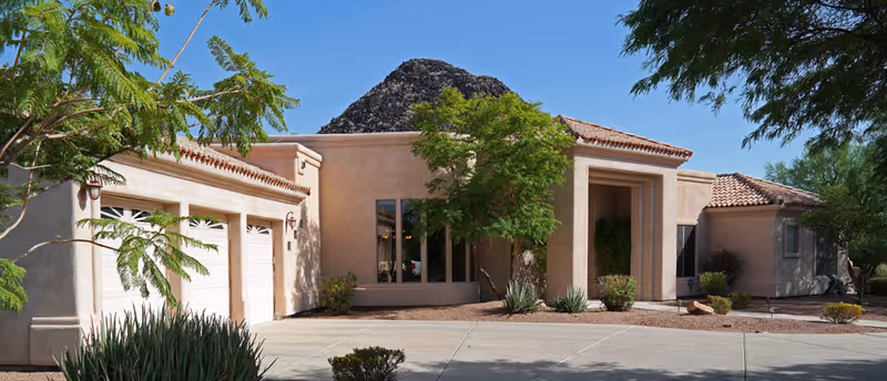 Single-story Southwestern-style stucco building with a three-car garage, front entrance columns, and desert landscaping under a clear blue sky.