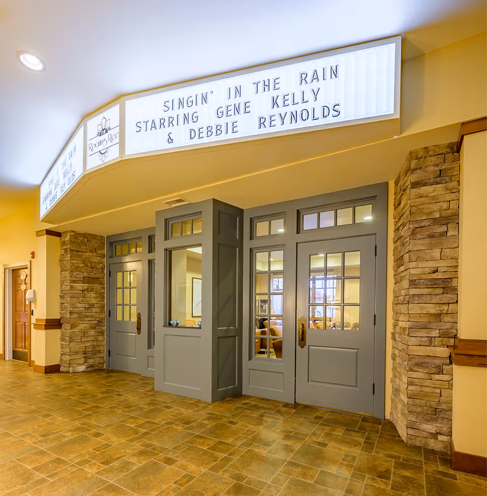 Interior view of a hallway in Creekside Assisted Living LLC featuring a set of gray double doors with glass panes and stone pillars on either side. Above the doors is a lighted marquee sign displaying the text 'SINGIN' IN THE RAIN STARRING GENE KELLY & DEBBIE REYNOLDS'. The floor is tiled with a brown and tan pattern, and the walls are painted yellow.