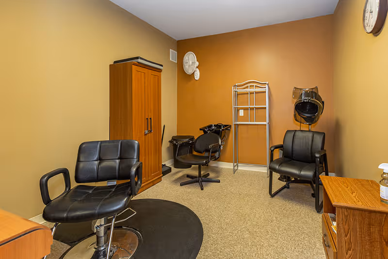 Interior of a small salon room with three black salon chairs, a wooden cabinet, a hair dryer hood, a wall-mounted fan, and a metal rack against tan and brown walls.