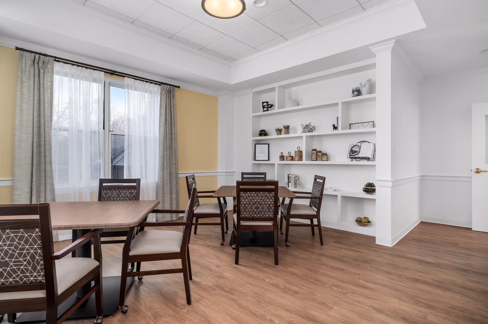 A bright and clean dining area with wooden tables and chairs featuring patterned backs and cushioned seats. The room has light yellow walls with white trim, a large window with sheer white curtains and gray drapes, and built-in white shelves decorated with small plants and decorative items. The floor is wood, and the ceiling has recessed lighting.