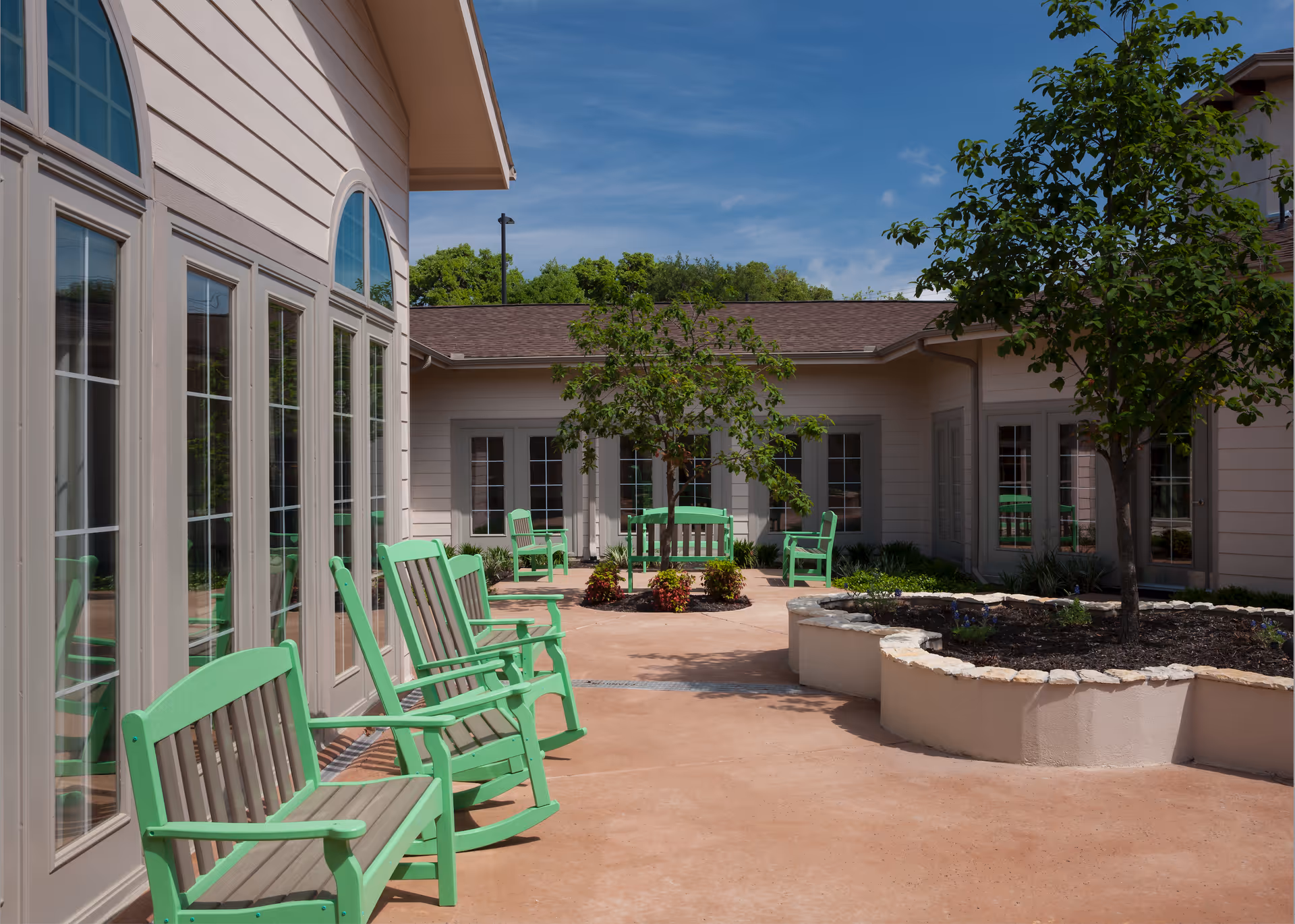Outdoor courtyard area at Franklin Park Boerne with green wooden rocking chairs and benches arranged around a paved patio. There are small trees and landscaped garden beds with plants, surrounded by beige building walls with multiple windows and doors under a clear blue sky.