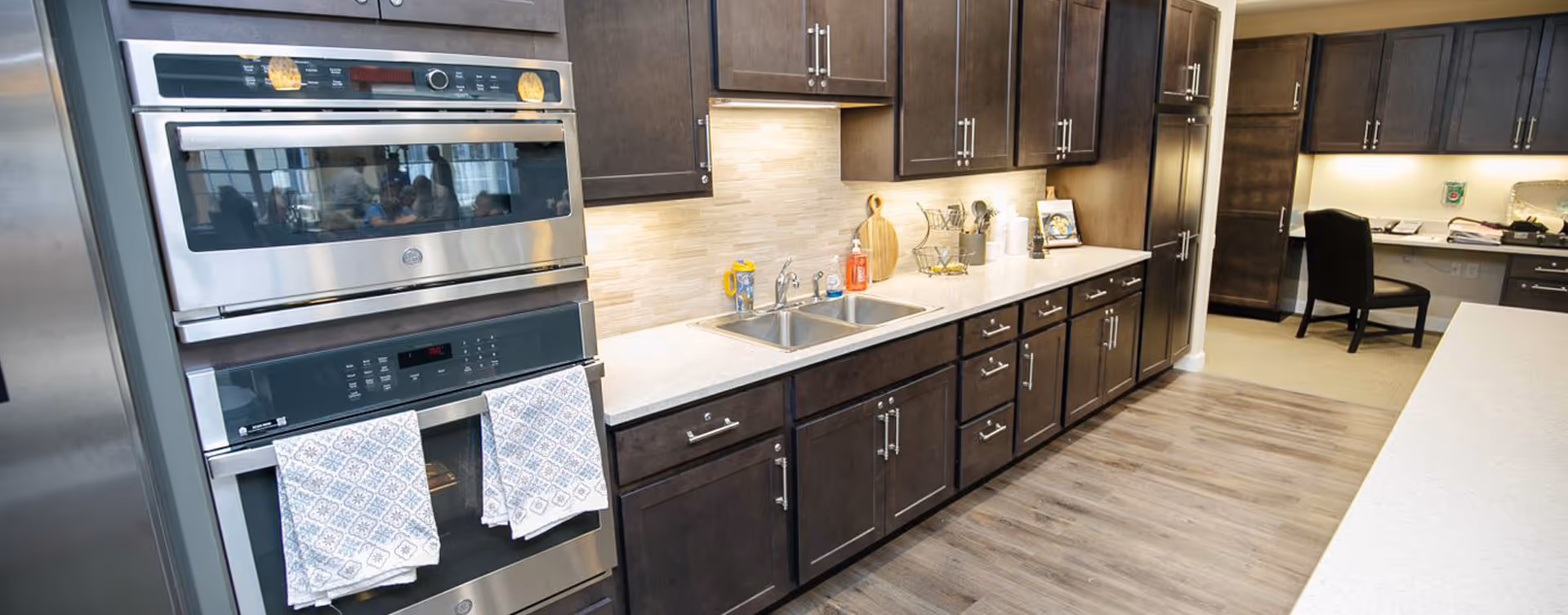 Modern kitchen with dark wood cabinets, stainless steel double oven, double sink, and light-colored countertops. The kitchen has under-cabinet lighting and various kitchen utensils and items on the counter. In the background, there is a desk area with a black chair and office supplies.