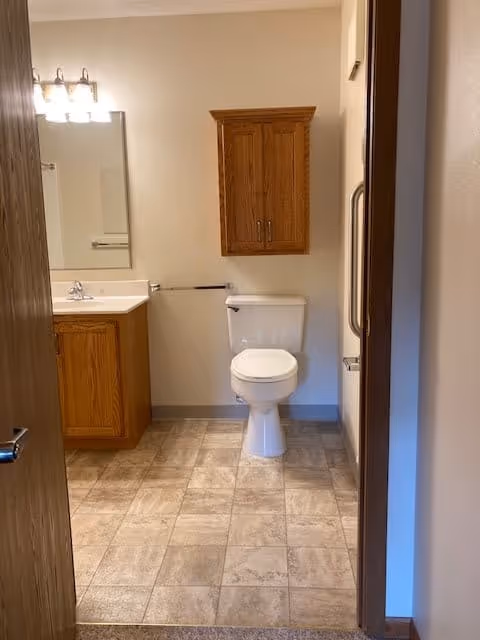 View into a bathroom with a white toilet centered against the back wall, a wooden cabinet mounted above the toilet, a wooden vanity with a sink and mirror on the left side, and beige tiled flooring.